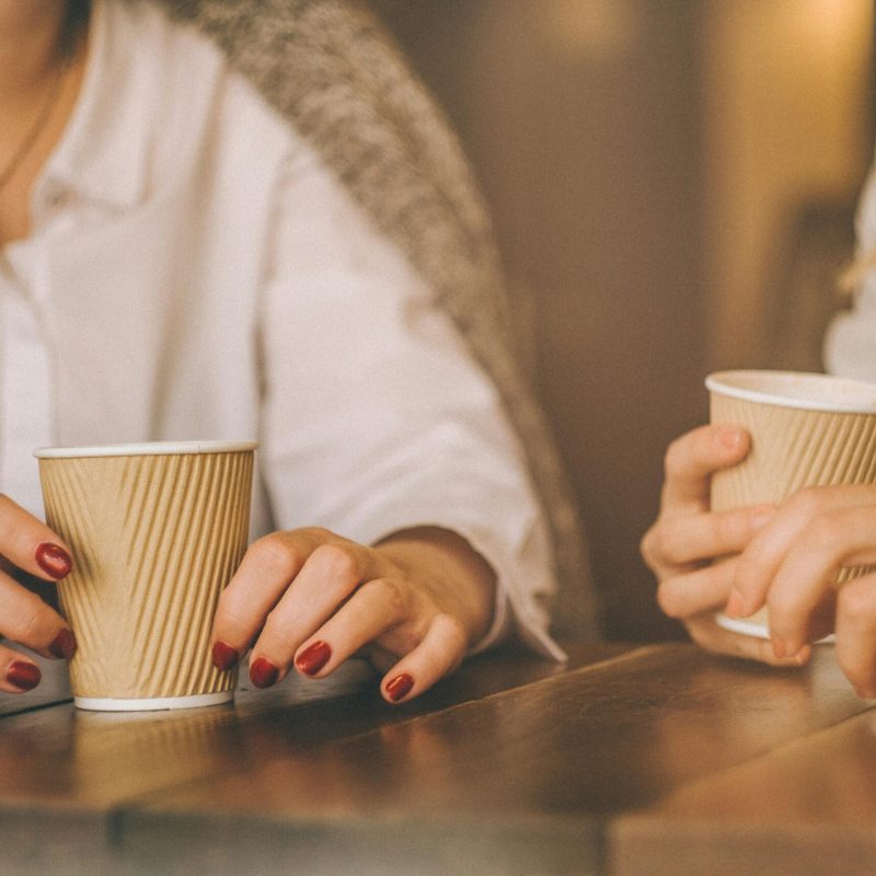 Two people holding paper coffee cups on a wooden table indoors, sharing a moment.